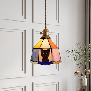 May include: A stained glass pendant lamp featuring a central black cat silhouette on a multi-colored panel background of yellow, orange, pink, and blue. The lamp has a brass-toned metal fixture and hangs from a twisted brown fabric-covered cord.