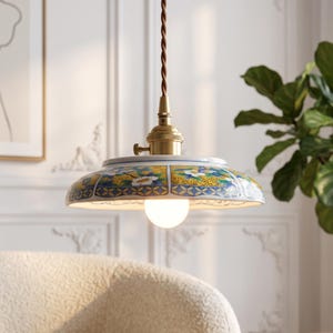 May include: A pendant light featuring a shallow, wide ceramic shade decorated with a blue, yellow, and white floral pattern. The fixture includes a brass-toned metal socket and a brown twisted fabric cord, suspended with a single round light bulb.