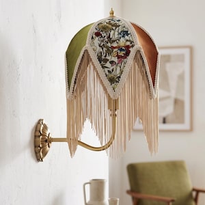 May include: A wall-mounted lamp featuring a multi-paneled shade with olive green and rust-colored velvet sections alternating with floral-patterned fabric panels. The shade is trimmed with cream-colored decorative braid and long fringe.