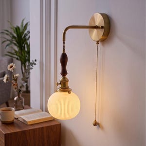 May include: A wall-mounted lamp featuring a circular brass base, a curved brass arm, and a dark wood accent piece. The light fixture includes a spherical, ribbed white glass shade and a brass pull chain with a small spherical end piece.