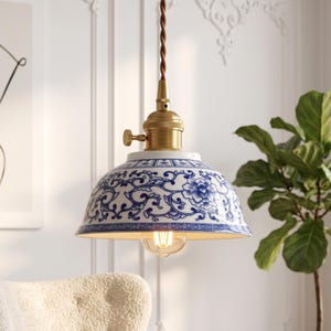 May include: A pendant light featuring a dome-shaped ceramic shade with a blue and white floral pattern. The fixture includes a brass-toned metal socket with a small side knob and a brown twisted fabric cord for suspension from the ceiling.