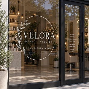 May include: A glass storefront window featuring a white circular decal with a leaf branch design and the text VELORA BEAUTY ATELIER SKIN BROWS LASHES. To the right, white text displays hours: MONDAY-FRIDAY 9AM-7PM, SATURDAY 10AM-5PM, SUNDAY CLOSED.