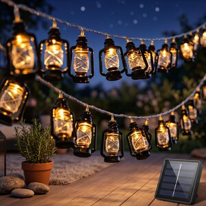 Può includere: Una catena di luci LED da esterno con mini lanterne nere e lampadine a luce calda. Le luci sono collegate da un cavo bianco e includono un'unità di ricarica solare separata di forma rettangolare e di colore nero.