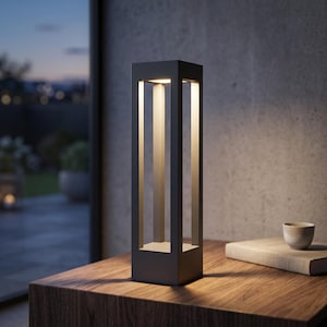 Pode incluir: Um candeeiro de mesa alto e retangular de cor cinzento-escuro com um design de estrutura aberta. O candeeiro apresenta uma base e um topo sólidos ligados por quatro pilares verticais, emitindo uma luz quente virada para baixo.