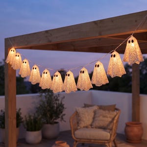 Può includere: Una catena luminosa con coperture all'uncinetto bianco a forma di fantasma appese a una struttura in legno. Ogni fantasma presenta due occhi circolari scuri ed è illuminato dall'interno da una luce calda, creando un effetto luminoso lungo il filo.
