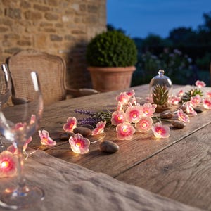 May include: A string of LED fairy lights featuring translucent pink flower-shaped covers with yellow centers. The lights are arranged across a wooden surface, interspersed with smooth grey river stones and sprigs of purple lavender.