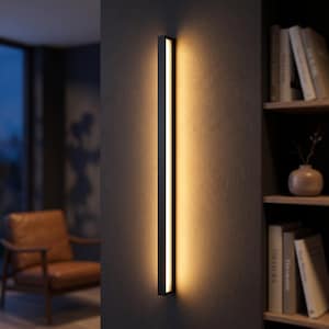 May include: A long, vertical, rectangular wall light fixture with a matte black frame and a central light strip that emits a warm, soft glow, casting a vertical beam of light onto the dark gray wall surface behind the fixture.