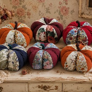 May include: Six fabric pumpkins arranged in two rows, featuring alternating segments of solid-coloured velvet and floral-patterned fabric. Each pumpkin is topped with a small, dark brown stem. The colours include orange, deep red, and navy blue.