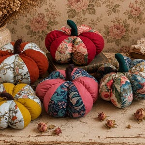 May include: A collection of five stuffed fabric pumpkins featuring alternating panels of solid-coloured velvet and floral-patterned cotton fabric. Each pumpkin is topped with a small, dark brown or dark green fabric stem, showcasing various colours including mustard yellow, deep red, pink, and blue.