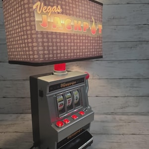 May include: A table lamp featuring a rectangular shade with a brown pattern of dollar signs and the text Vegas JACKPOT. The base is a silver-colored slot machine replica with three spinning reels, red illuminated buttons, and a side lever.