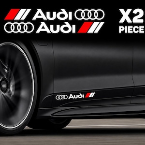 Peut inclure: Un ensemble de deux autocollants en vinyle blanc présentant le logo et le texte Audi avec un motif de rayures inclinées rouges et blanches. Les autocollants sont appliqués sur le panneau latéral inférieur d'une voiture noire.