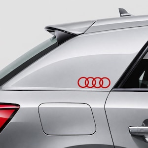 May include: A silver car features a red decal of the four interlocking rings logo applied to the rear side window. The vehicle has a sleek body design with a visible fuel door, a door handle, and a black spoiler on the upper rear section.