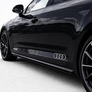 Peut inclure: Une berline Audi noire vue de profil, arborant un autocollant argenté avec le logo aux quatre anneaux sur le panneau inférieur de la porte arrière. Le véhicule présente des jantes en alliage sombres à rayons multiples et un rétroviseur noir.