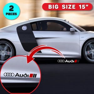 May include: A set of two 15-inch Audi car decals featuring the brand logo and name in black, followed by three vertical stripes in red, black, and grey. The stickers are shown applied to the side of a silver sports car with a V10 engine badge.