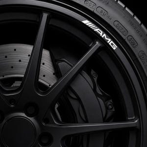 May include: A matte black automotive wheel featuring thin spokes and a white AMG logo printed on the rim edge. The wheel is paired with a black tyre showing DOT markings and a metallic brake rotor with circular ventilation holes visible behind.
