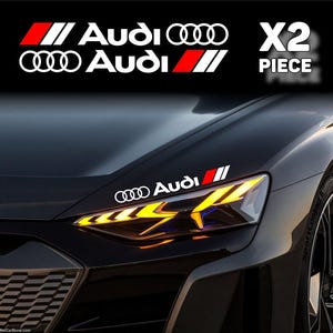 Può includere: Un set di due adesivi in vinile bianco con il logo e il nome del marchio Audi, accompagnati da una grafica a strisce oblique rosse e bianche. Gli adesivi sono applicati sul cofano di un'auto grigio scuro sopra il faro anteriore.