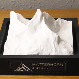 May include: A white 3D topographic model of the Matterhorn mountain peak set inside a square black frame. A rectangular plaque on the front of the frame displays a small mountain icon, the text MATTERHORN, and the elevation of 4,478 m.