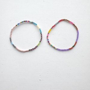 May include: Two circular bracelets made of small, multicolored seed beads. The left bracelet features a mix of pink, blue, grey, and green beads. The right bracelet displays a vibrant sequence of red, yellow, purple, orange, and blue beads.