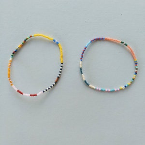 May include: Two colorful beaded bracelets. One bracelet is yellow, blue, red, and white. The other bracelet is pink, blue, white, and green.