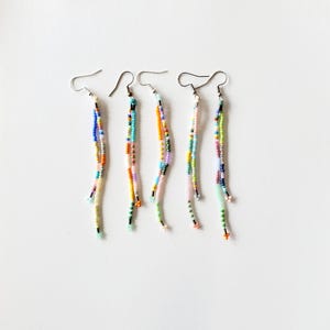 May include: Four pairs of beaded dangle earrings. Each earring features a silver hook and a long strand of colorful beads in various hues, including blue, orange, green, and pink. The earrings are arranged against a white background.