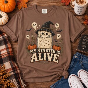May include: A brown short-sleeve t-shirt featuring a graphic of a sourdough starter in a glass jar wearing a witch hat, surrounded by ghosts, bats, wheat stalks, and pumpkins, with the text MY STARTER IS ALIVE printed in large letters.