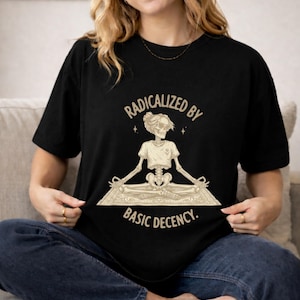 Könnte beinhalten: Ein schwarzes Kurzarm-T-Shirt mit einer beigen Grafik eines Skeletts, das in der Lotus-Yoga-Position auf einer Matte sitzt. Das Skelett trägt eine Sonnenbrille und einen Dutt. Der Text lautet RADICALIZED BY BASIC DECENCY in beiger Schrift.