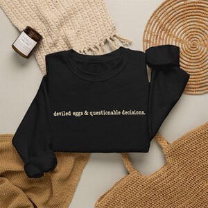 May include: A black long-sleeve crewneck sweatshirt featuring the text "deviled eggs & questionable decisions." printed in a light beige, typewriter-style font across the center of the chest. The garment has a ribbed collar, cuffs, and hem.