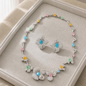 Könnte beinhalten: Eine silberfarbene Halskette und passende Ohrringe mit Blumenmotiven, besetzt mit klaren Pavé-Steinen und akzentuiert durch mehrfarbige, oval geschliffene Edelsteine in Blau, Pink, Gelb und Grün, angeordnet an einer feinen Gliederkette.