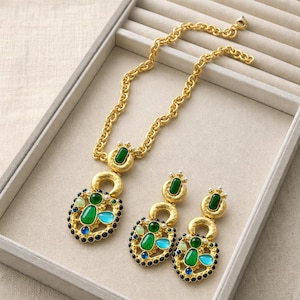 Könnte beinhalten: Eine goldfarbene Halskette und ein passendes Paar Ohrhänger mit strukturierter Metalloberfläche. Jedes Schmuckstück ist mit grünen, türkisfarbenen und blauen Cabochon-Steinen, kleinen weißen Kunstperlen und einem Rand aus winzigen dunkelblauen Perlen verziert.