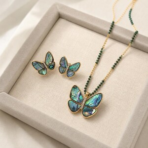 Könnte beinhalten: Ein Schmuckset bestehend aus einer Halskette und einem Paar Ohrsteckern, jeweils mit Schmetterlingsdesign, Flügeln aus irisierendem Abalone-Muschelmaterial, goldfarbenen Metallrändern und einer zentralen Reihe kleiner klarer Kristalle.