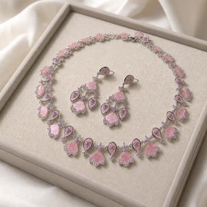 Könnte beinhalten: Ein Schmuckset bestehend aus einer Halskette und passenden Ohrhängern. Die Stücke verfügen über silberfarbene Metallfassungen, klare Pavé-Kristalle und hellrosa, fächerförmige Steinakzente in einem symmetrischen Muster.
