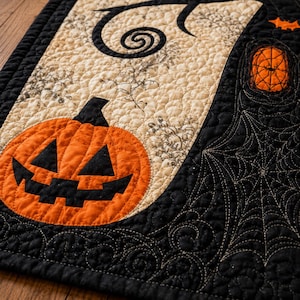 May include: A quilted fabric runner featuring a large orange jack-o'-lantern with a black face on a cream background with black swirling patterns. The right side is black with an orange spiderweb design and a small orange pumpkin motif.
