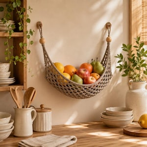 Puede incluir: Una hamaca para frutas de macramé beige suspendida por dos ganchos de madera, que contiene una variedad de productos frescos que incluyen manzanas rojas, peras verdes y cítricos naranjas. La red tejida presenta un patrón de rombos y cuentas de madera.