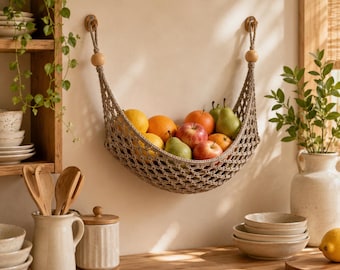 Puede incluir: Una hamaca para frutas de macramé beige suspendida por dos ganchos de madera, que contiene una variedad de productos frescos que incluyen manzanas rojas, peras verdes y cítricos naranjas. La red tejida presenta un patrón de rombos y cuentas de madera.