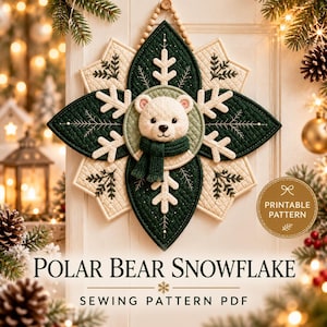 Pode incluir: Uma tapeçaria acolchoada com o rosto de um urso polar ao centro usando um cachecol verde-escuro, rodeado por um design em forma de estrela de pétalas de tecido verde-escuro e creme com detalhes de flocos de neve bordados e um laço de contas.