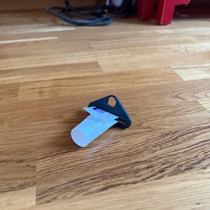Op de afbeelding: Een kleine witte plastic tube met een witte dop, geplaatst in een zwarte driehoekige plastic tube-uitknijper. De tube is voorzien van een blauw-wit etiket met zichtbare tekst. Het hulpmiddel is ontworpen om de inhoud uit de tube te drukken.