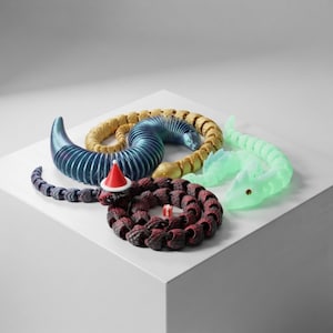 Puede incluir: Cuatro figuras de serpiente articuladas de varios colores y texturas. Una es azul metálico, una dorada, una verde claro translúcido y una roja oscura con un pequeño gorro de Papá Noel rojo con borde blanco y un diminuto regalo.