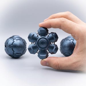 Peut inclure: Un fidget spinner métallique gris foncé en forme d'étoile à six branches avec des segments arrondis et un roulement central circulaire. Deux objets sphériques supplémentaires aux surfaces segmentées gris foncé assorties sont présents.