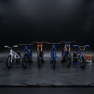 Pode incluir: Uma coleção de seis modelos de bicicletas em miniatura dispostos em fila. As bicicletas apresentam várias cores de quadro, incluindo branco, azul, vermelho e acabamentos iridescentes. Cada modelo inclui guiadores, rodas e quadros detalhados.