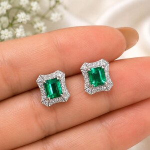 May include: A pair of stud earrings featuring emerald-cut green gemstones set in silver-toned metal. Each central green stone is surrounded by a rectangular halo of small, clear, sparkling white stones, creating a classic and elegant appearance.