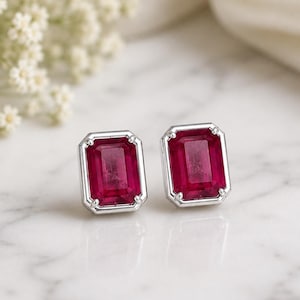 May include: A pair of stud earrings featuring rectangular, emerald-cut deep red gemstones set in polished silver-toned metal. Each stone is secured by four prongs, creating a clean, geometric look with a vibrant contrast between the red and silver.