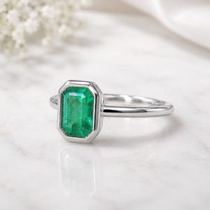 Peut inclure: Une bague argentée ornée d'une pierre précieuse verte de taille émeraude rectangulaire, fixée dans un serti clos lisse. L'anneau est poli et fin, présentant un design épuré qui met en valeur la couleur verte vive de la pierre centrale.
