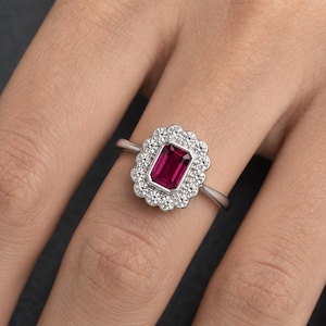 May include: A silver-toned ring featuring a central rectangular emerald-cut deep red gemstone surrounded by a scalloped halo of small, clear, round-cut stones. The band is thin and polished, creating a refined look with a vibrant contrast.