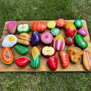 May include: A collection of smooth river stones hand-painted with colourful designs of various foods including fruits, vegetables, a hamburger, a hot dog, a slice of pizza, a doughnut, an ice cream cone, popcorn, and a fried egg on a wooden board.