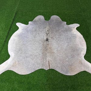 May include: A natural cowhide rug featuring a mottled pattern of grey, white, and light brown tones. The hide has an irregular, organic shape with a textured surface and a central dark marking, displaying the authentic hair and hide characteristics.