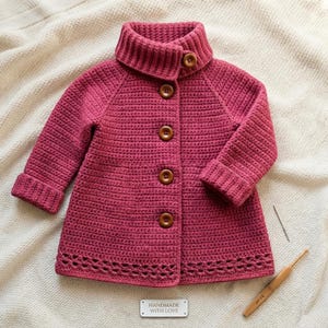 Può includere: Un cardigan all'uncinetto color magenta con collo alto, maniche lunghe con polsini risvoltati e cinque bottoni in legno marrone sul davanti. L'orlo presenta un motivo traforato decorativo. Una piccola etichetta rettangolare riporta HANDMADE WITH LOVE.