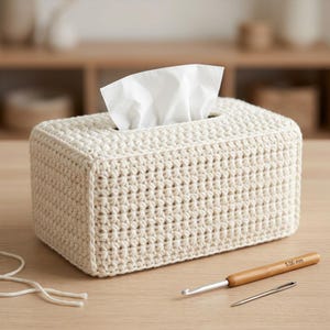 May include: A rectangular tissue box cover made of cream-colored yarn in a dense crochet stitch pattern. A white tissue protrudes from the top opening. A wooden crochet hook labeled 5.00 mm and a metal sewing needle lie on the surface nearby.