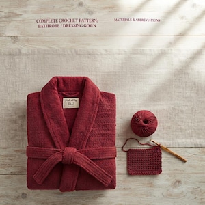 Puede incluir: Un albornoz de color rojo intenso con cuello de chal y cinturón anudado, con una etiqueta que dice Handmade by JKD. Junto a él hay un ovillo de lana a juego, una pequeña muestra rectangular de ganchillo y un ganchillo de madera.