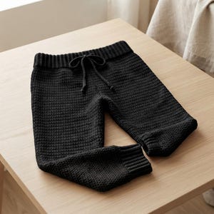 May include: Solid black knit trousers featuring a textured stitch pattern, a ribbed elastic waistband with a matching drawstring tie, and ribbed cuffs at the ankles. The garment is constructed with a consistent dark yarn throughout the entire design.