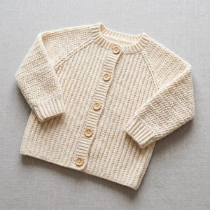 May include: A cream-colored long-sleeve cardigan made of thick, textured crochet knit fabric. The garment features a round neckline, ribbed cuffs, and a vertical front placket secured with five round, light brown wooden buttons down the center.